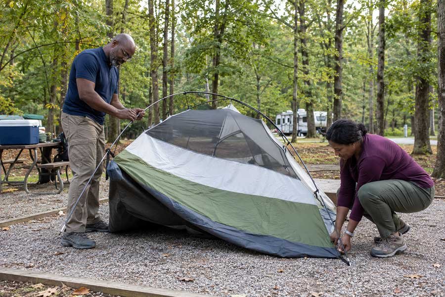 Beginner setting up a tent at a campground with trees and a picnic table nearby